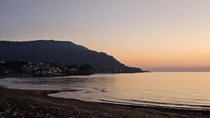 Escena costanera amb una platja, mar tranquil·la i un turó amb edificis al fons.