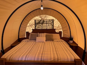 A tented room with a bed, wooden headboard, and a hanging light fixture. - Charming Conestoga Wagon Rental for Unforgettable Glamping in Mineral Wells, Texas (Mineral Wells)