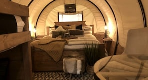 A cozy, arched room with a bed, a chair, and a small bench. - Charming Conestoga Wagon Rental for Unforgettable Glamping in Mineral Wells, Texas (Mineral Wells)