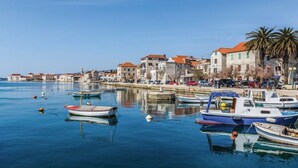Un pueblo costero con barcos amarrados a lo largo del muelle, edificios de piedra con tejados rojos y un cielo azul despejado.