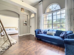 Ein Zimmer mit blauem Sofa, Schaukelstuhl, Bett und einem großen Fenster mit Vorhängen.