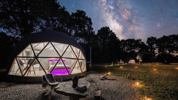 A geodesic dome structure with a bed and seating area, surrounded by a gravel path and trees, under a starry night sky.
