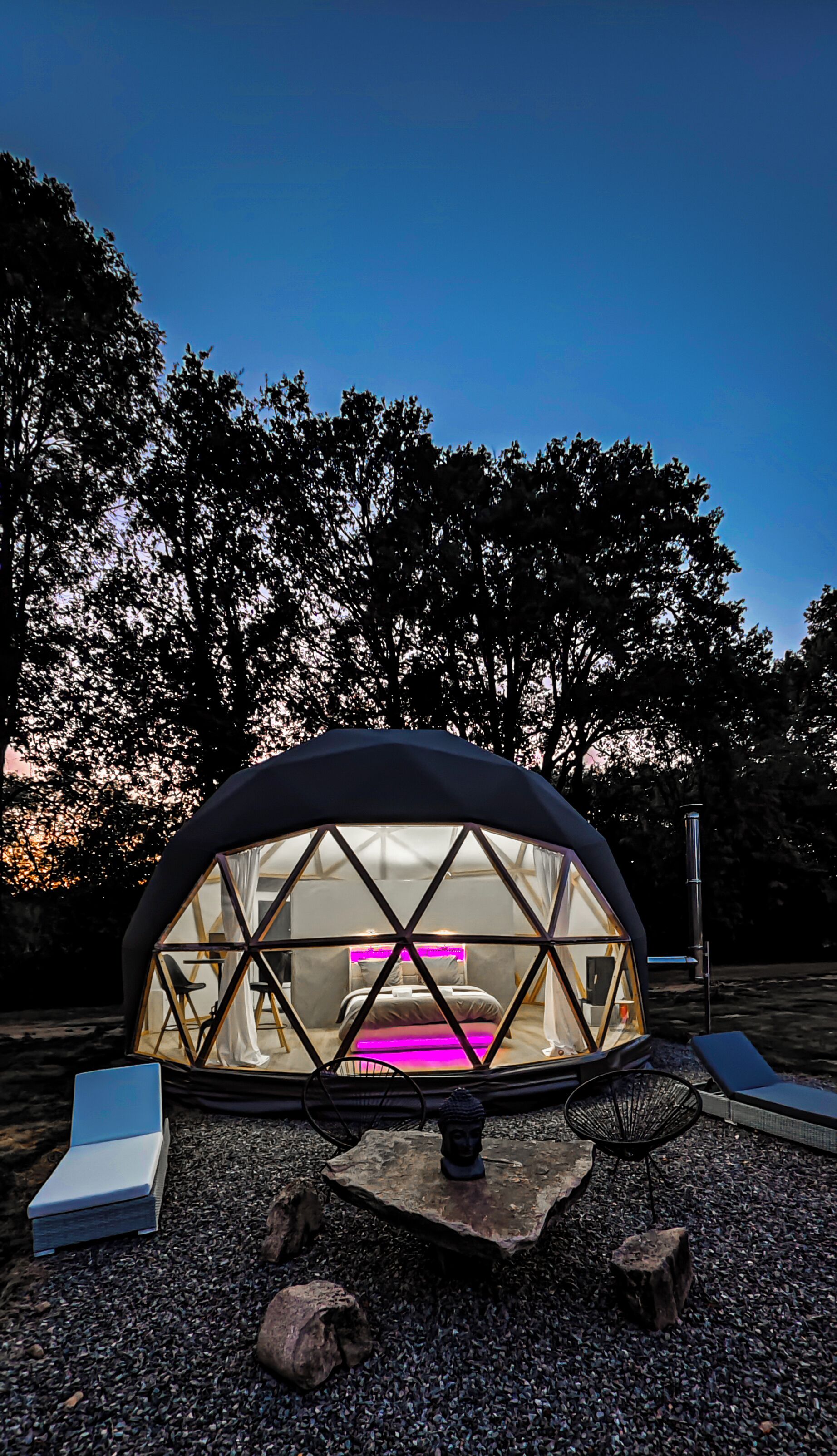 Een geodetische koepelstructuur met een bed en zithoek binnenin, omgeven door bomen en een grindpad.