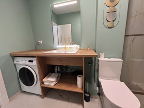 A bathroom with a washing machine, a toilet, a sink with a mirror, and a shelf with towels.