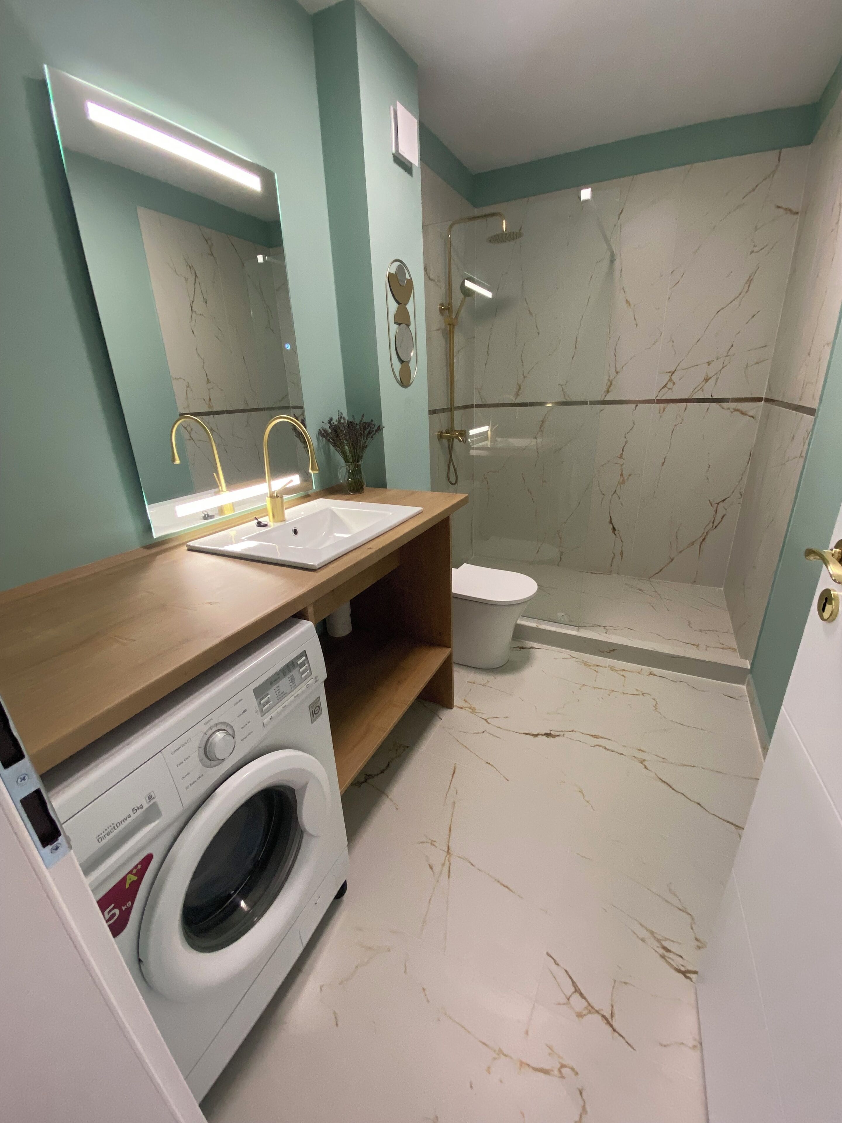 Ein modernes Badezimmer mit Marmorboden, einer Waschmaschine, einem Waschbecken mit goldenen Armaturen und einer Duschkabine.