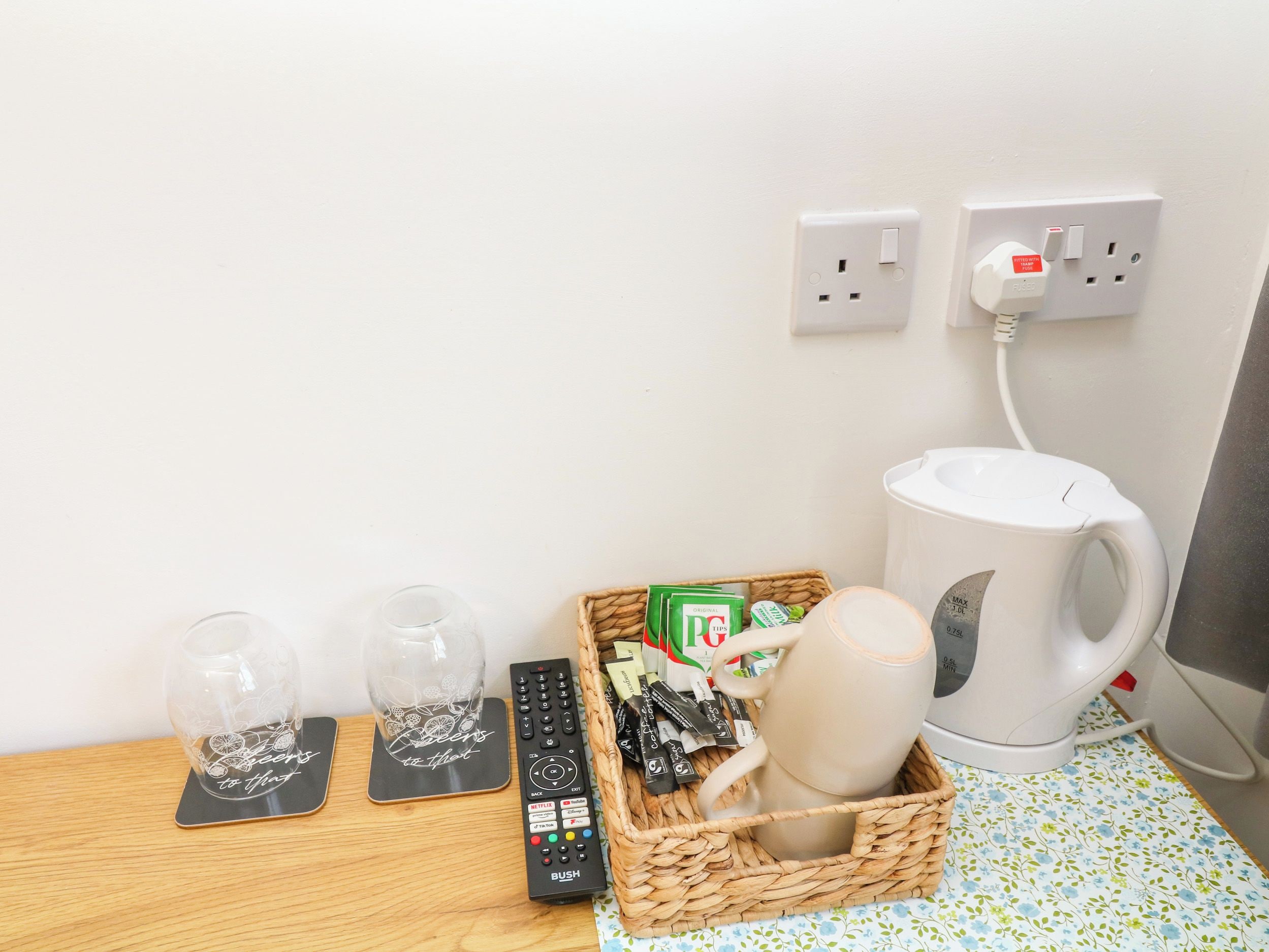 A room with a wooden table, a coffee maker, a basket with remote controls and a mug, and a wall socket.