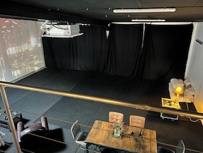 A room with a stage, black curtains, a table with chairs, a projector, and a lamp.