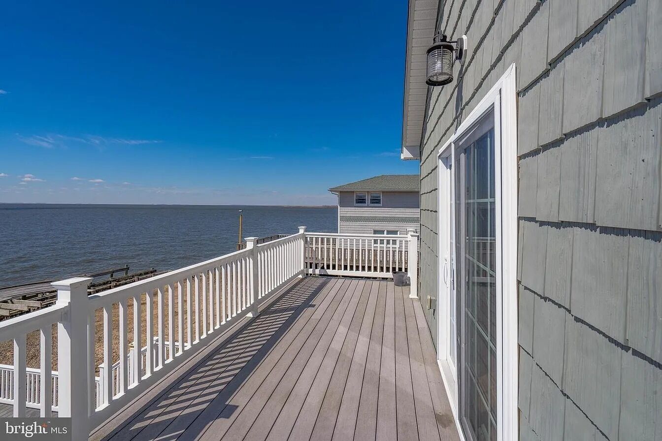 A wooden deck with a white railing, a building with a gray siding, and a view of the water.