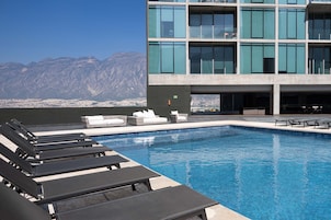 Una piscina con sillones de descanso, un edificio con amplias ventanas y montañas al fondo.