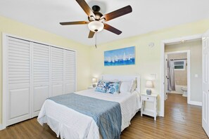 A bedroom with a bed, a ceiling fan, a painting, a nightstand, and a bathroom door. - Low Key Tiki (Cape Coral)