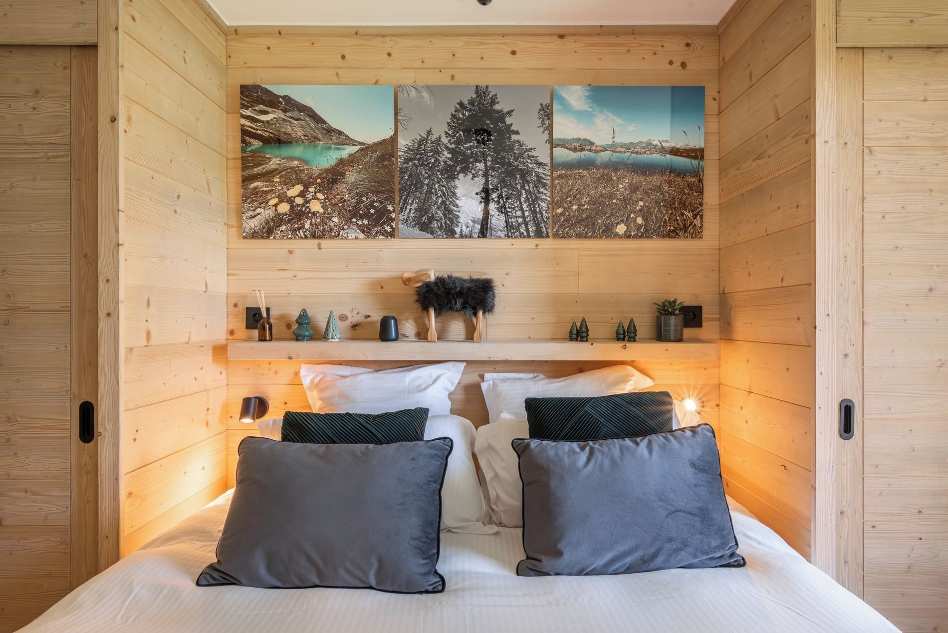 A wooden interior with a bed, pillows, a shelf with decorative items, and a wall with three paintings.