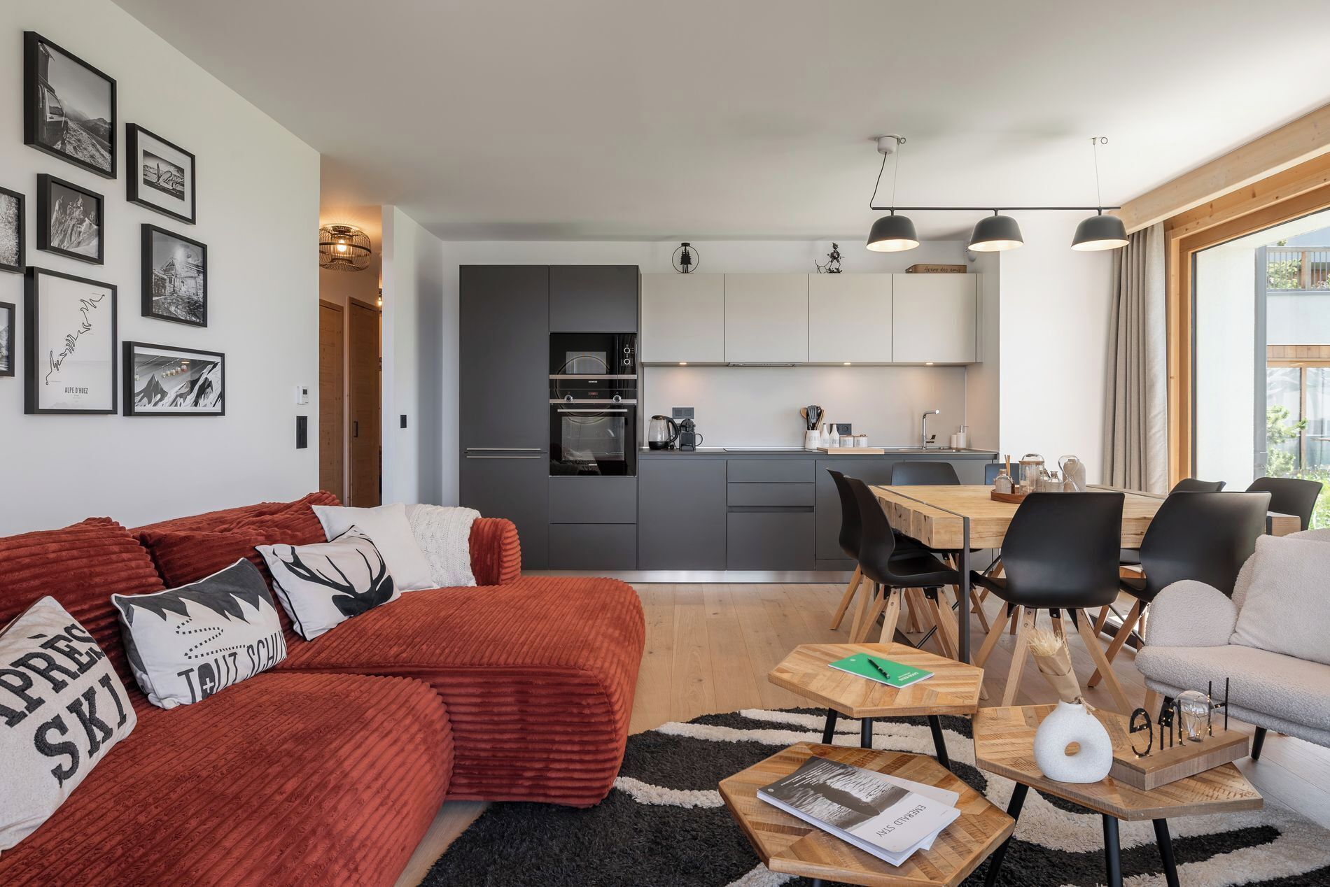 Ein modernes Wohnzimmer mit einem roten Sofa, einem Essbereich mit Holztisch und einer Küche mit grauen Schränken.