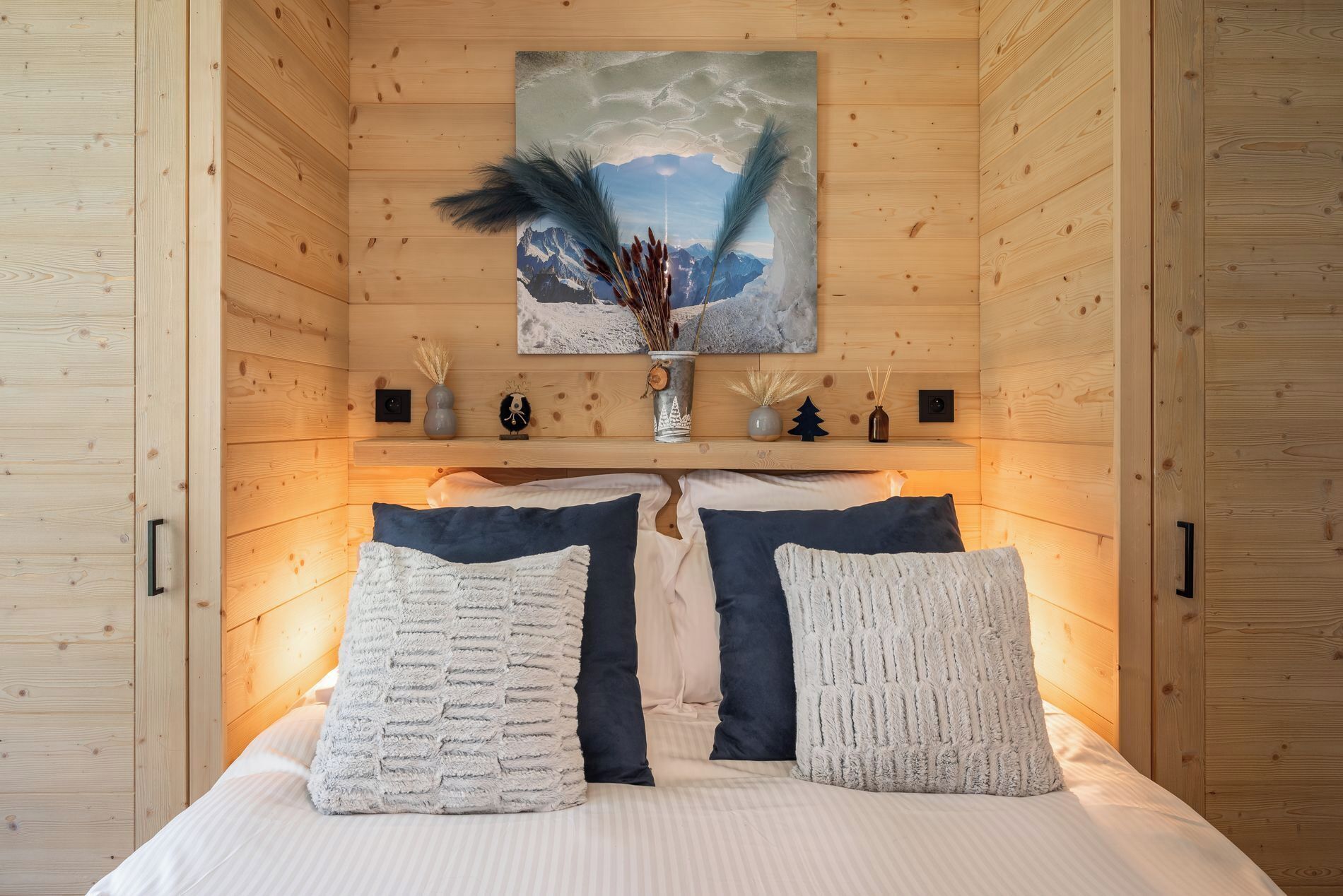 Een gezellig houten kamer met een bed, kussens, een schilderij en een plank met decoratieve spullen.