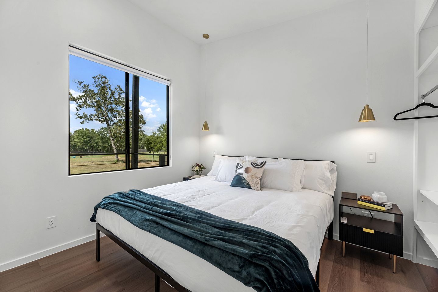 A modern bedroom with a large window, a bed with white linens, a bedside table with books, and a hanging light fixture.