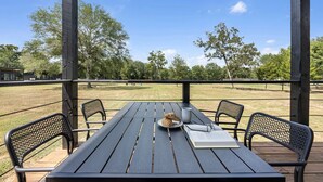 An outdoor wooden table with chairs, a dog, and a book on a deck with a view of a grassy field and trees. - Modern Farmhouse Tiny Home Resort with Goats & Pool near Houston, Hempstead Texas (Hempstead)