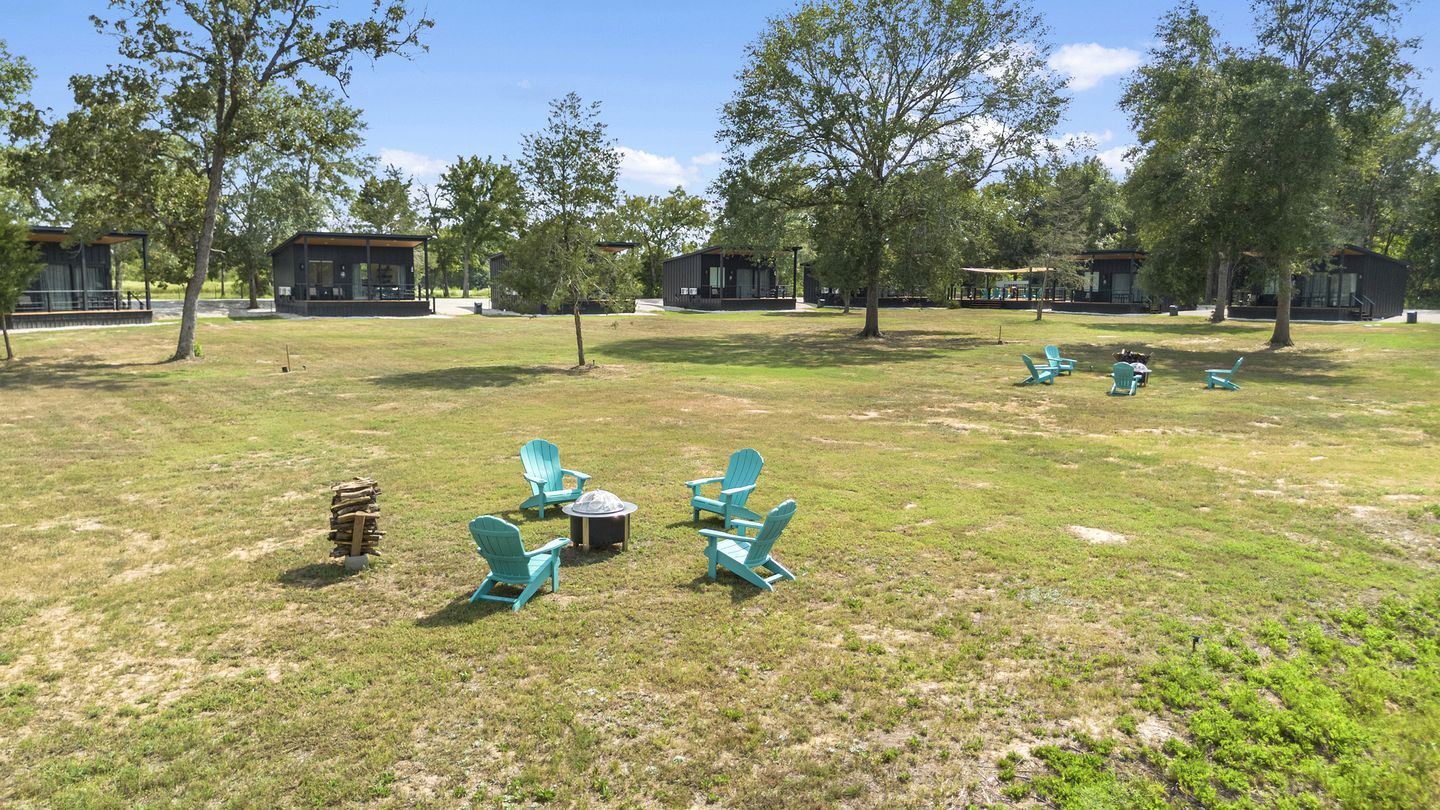 A grassy outdoor area with several Adirondack chairs arranged in a circle around a fire pit, surrounded by trees and small cabins.