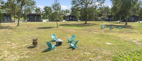 A grassy outdoor area with several Adirondack chairs arranged in a circle around a fire pit, surrounded by trees and small cabins.