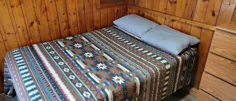 A single bed with a patterned blanket and a light blue pillow in a room with wooden walls and a window.
