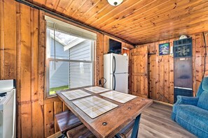 A cabin interior with a wooden ceiling, a dining table with placemats, a refrigerator, a couch, and a window with blinds. - Lakefront Cottage with Fire Pit, Beach & Pet Friendly in Houghton Lake, Michigan (Houghton Lake)