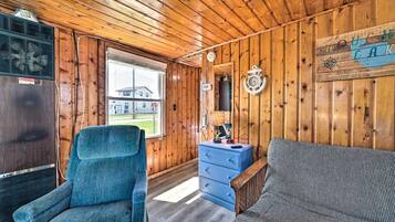 A cabin interior with a wooden ceiling, a blue chair, a sofa, a dresser, and a window with a view of a house.