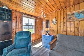 A cabin interior with a wooden ceiling, a blue chair, a sofa, a dresser, and a window with a view of a house. - Lakefront Cottage with Fire Pit, Beach & Pet Friendly in Houghton Lake, Michigan (Houghton Lake)