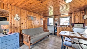 A wooden cabin interior with a sofa, a dining table, a kitchen area, and a clock on the wall.