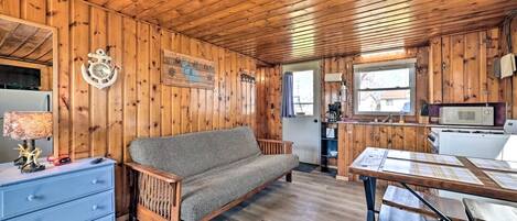 Interior de una cabaña de madera con sofá, una mesa de comedor, una zona de cocina y un reloj en la pared.