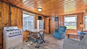 A cabin interior with a wooden floor, a stove, a refrigerator, a dining table with benches, a blue chair, and a sofa.
