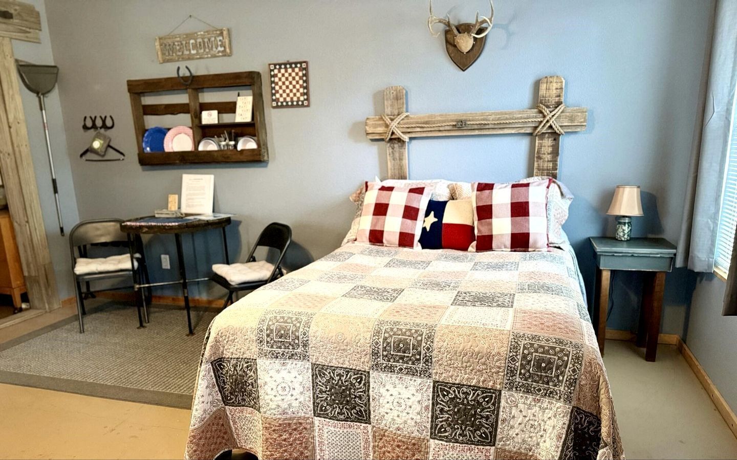 A bedroom with a quilted bedspread, a desk with a chair, a wooden cross decoration, and a welcome sign on the wall.