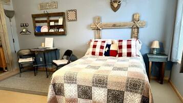 A bedroom with a quilted bedspread, a desk with a chair, a wooden cross decoration, and a welcome sign on the wall.