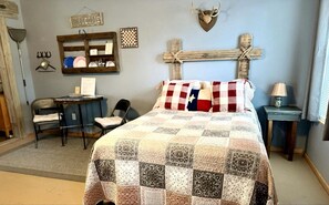 Ein Schlafzimmer mit gesteppter Bettdecke, einem Schreibtisch mit Stuhl, einer Holzkreuz-Dekoration und einem Willkommensschild an der Wand.