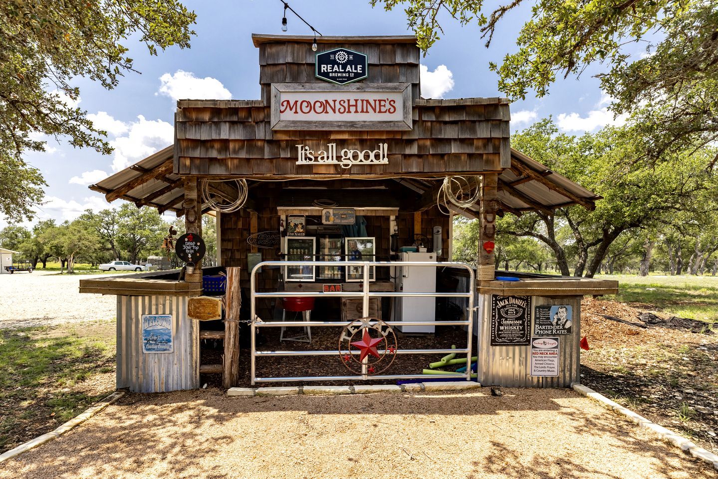 A rustic outdoor bar named 'MOONSHINE'S' with a sign stating 'It's all good!' and a 'BAR' sign on the door.