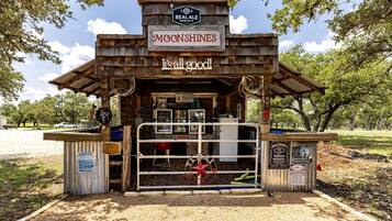 A rustic outdoor bar named 'MOONSHINE'S' with a sign stating 'It's all good!' and a 'BAR' sign on the door.