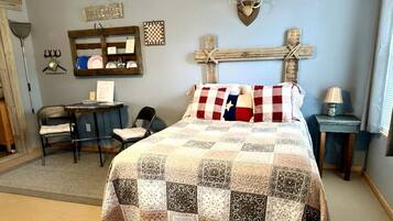 A bedroom with a quilted bedspread, a desk with a chair, a wooden cross decoration, and a welcome sign on the wall.