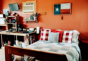 Een slaapkamer met een bed, een bureau met een computer, een plank met keukenbenodigdheden, en een Texas vlag.