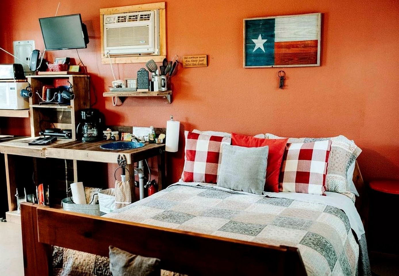 A bedroom with a bed, a desk with a computer, a shelf with kitchen items, and a Texas flag.