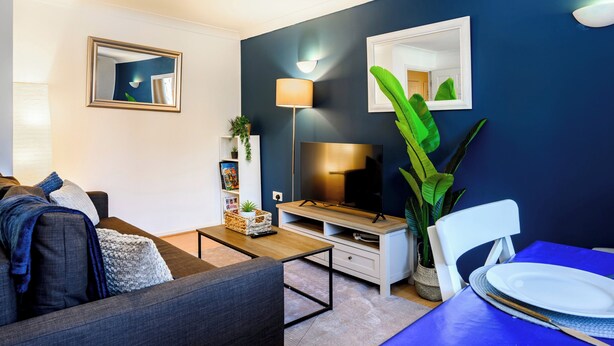 Ett modernt vardagsrum med en mörkblå accentvägg, en platt-TV, ett soffbord, en soffa med kuddar, ett matbord med tallrikar och en stor växt.