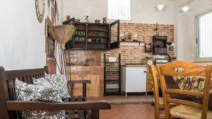 A cozy living area with a wooden bench, a dining table with chairs, a kitchen with a brick wall, and a window providing natural light. - Il Casolare Siciliano (Palmeri - Ferrante)