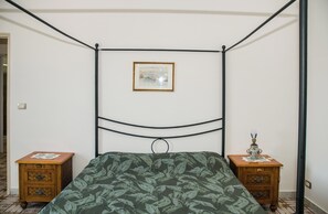 Ein Bett mit Himmel, dessen Bettdecke ein Grün-Blatt-Muster aufweist, zwei Nachttische aus Holz und ein gerahmtes Bild an der Wand.