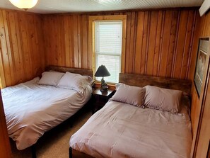 A room with two beds, a window with blinds, a nightstand with a lamp, and wooden paneling.