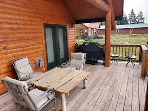 Una terraza de madera con una mesa, sillas, una parrilla y un área cubierta con vista a una casa al fondo.
