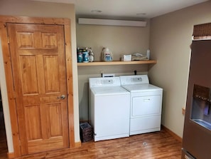 Un lavadero con puerta de madera, dos lavadoras blancas, un estante con productos de limpieza y un dispensador de agua.