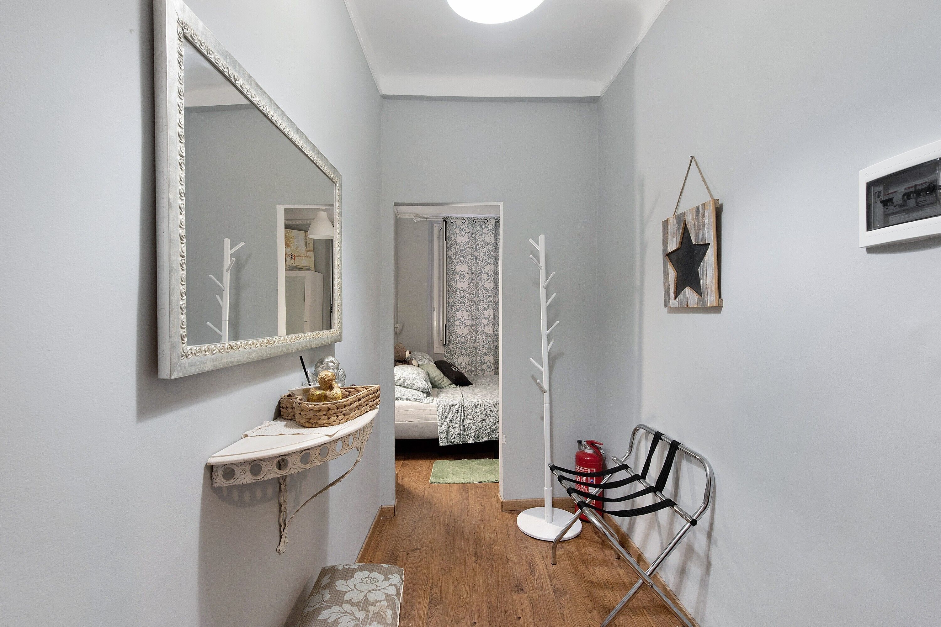 A hallway with a mirror, a coat rack, a bed with patterned bedding, and a small table with a basket.