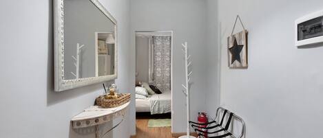 Een gang met een spiegel, een kapstok, een bed met behang en een klein tafeltje met een mandje.