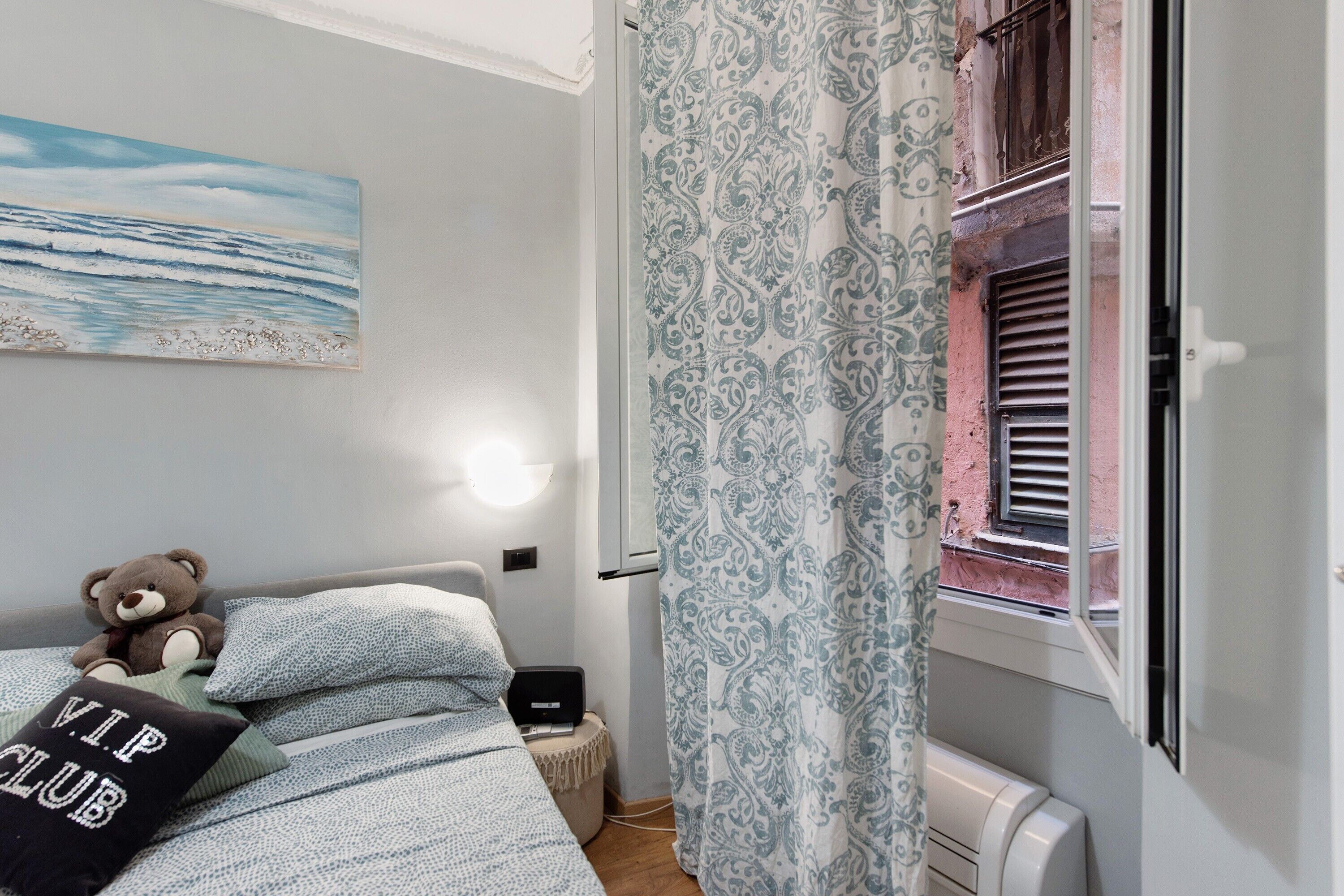 A bedroom with a bed, a teddy bear, a pillow with text, a painting, a patterned curtain, and a window with blinds.