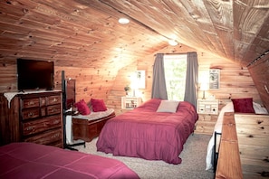 A cozy attic bedroom with a wooden ceiling, a bed with a red bedspread, a dresser, a television, and a window with curtains.