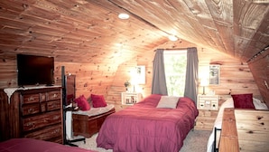 Une chambre confortable située dans les combles, avec un plafond en bois, un lit recouvert d’une couverture rouge, une commode, un téléviseur et une fenêtre avec des rideaux.