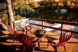 A wooden deck with Adirondack chairs and a fire pit, overlooking a lake with boats and houses in the background.