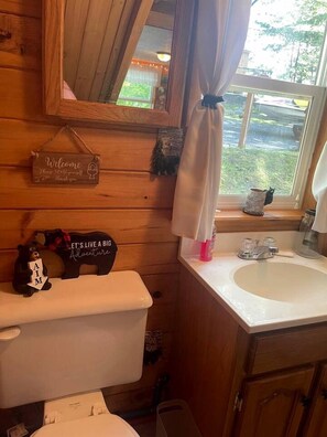 A wooden cabin bathroom with a toilet, sink, and window with curtains.
