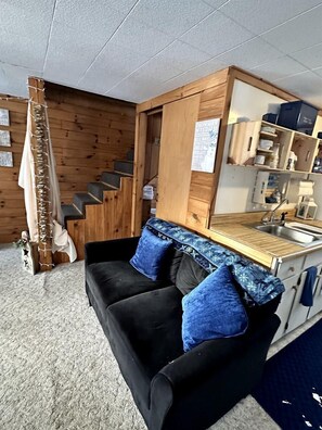 A compact living space with a black sofa, blue cushions, a wooden staircase, a kitchen area, and a small dining table.